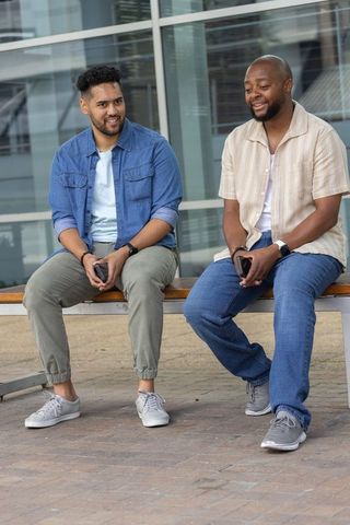African american men chatting on bench outside modern glass building, friendly chat