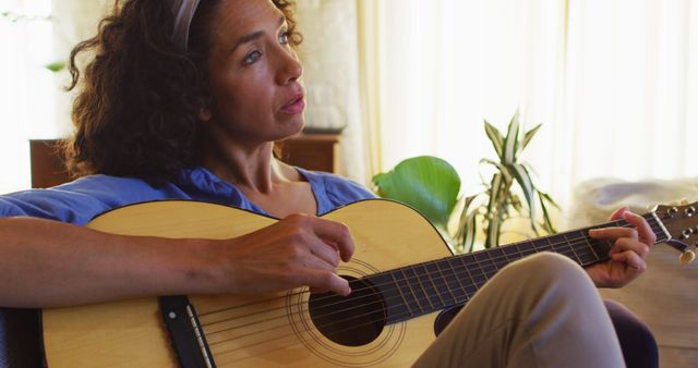 Woman sitting on sofa playing acoustic guitar in indoor setting. Image ideal for use in content related to music, relaxation, hobbies, creativity and home settings.