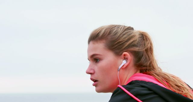 A young Caucasian woman is jogging outdoors, wearing earphones, with copy space. Her focused expression and sportswear suggest a commitment to fitness and a healthy lifestyle.