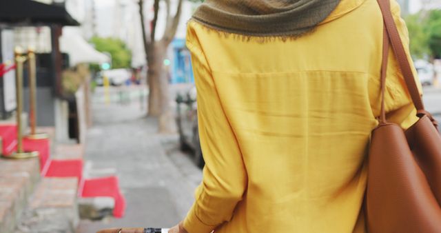 Rear midsection of woman yellow shirt with leather bag walking in city street. City living and modern urban lifestyle.