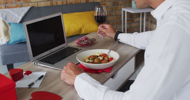 Man enjoying a virtual dinner date at home, eating pasta and drinking red wine while interacting with someone via a laptop. This image is great for use in articles or ads related to remote relationships, online connections, modern romance, or lifestyle blogs focusing on using technology to stay connected.