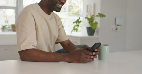 Smiling man leaning on kitchen counter holding smartphone and mug in bright modern home