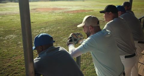 Diverse baseball teammates leaning on dugout rail watching game holding leather glove