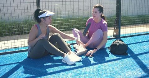Two women chatting and stretching on blue pickleball court, holding paddle and phone
