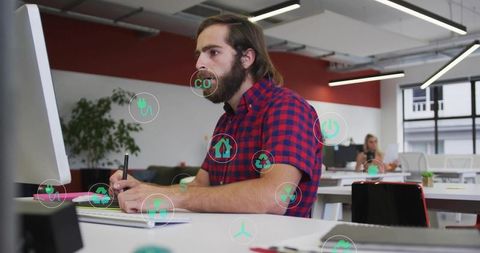 Bearded man writing at computer with green eco icons overlay in open office