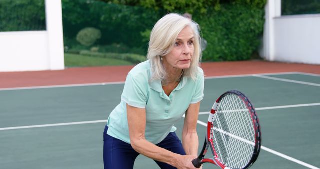 Senior woman holding a tennis racket on a tennis court, preparing to play. Ideal for fitness and health-related content, promoting active lifestyle among older adults, and sports activities for seniors.