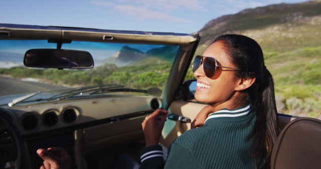 Woman wearing sunglasses smiling while seated in convertible car during road trip. She is enjoying the scenery and fresh air, with a picturesque view of the countryside in background. Ideal for concepts related to travel, adventure, relaxation, freedom, summer activities, and lifestyle promotions.