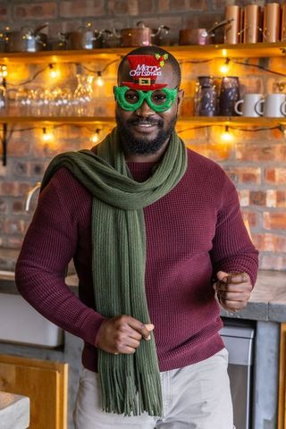 African american man wearing merry christmas glasses and scarf in cozy rustic kitchen