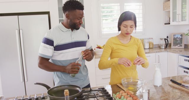 Couple preparing a meal in a bright, modern kitchen. Man holding seasoning bottle while woman cracks an egg into a bowl. Suitable for projects highlighting home cooking, togetherness, multicultural relationships, modern lifestyles, culinary activities.