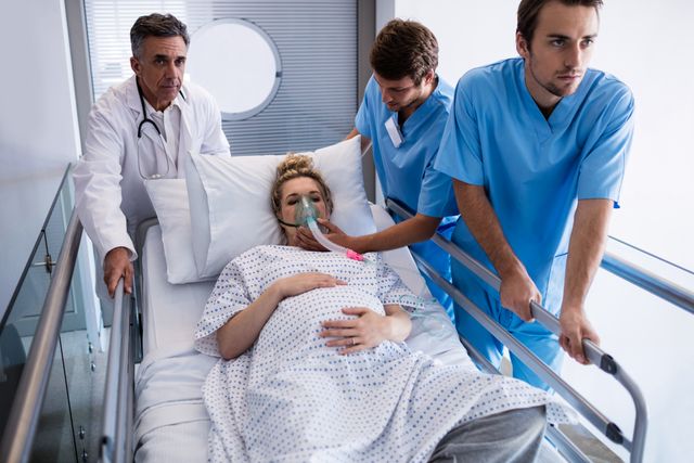 Medical team rushing a pregnant woman on a stretcher to the operation theatre in a hospital. The woman is wearing a hospital gown and an oxygen mask, indicating an emergency situation. This image can be used for healthcare, emergency medical services, maternity care, and hospital-related content.