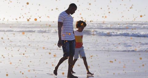 Walking father and son on sunlit beach with golden confetti and footprints