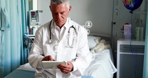Senior Doctor Reviewing Patient Records on Tablet in Hospital Room with Teddy Bear