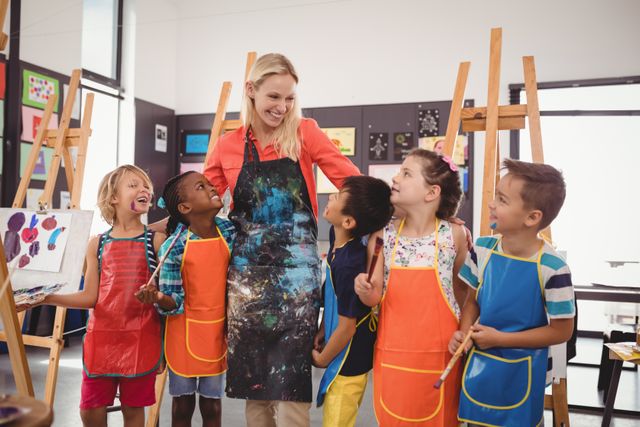 Teacher engaging with diverse group of children in art class. Kids wearing aprons, holding paintbrushes, and smiling. Ideal for educational content, school brochures, art programs, and diversity in education promotions.