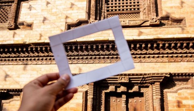 Hand holding paper frame framing ornate carved brick facade on sunlit historic street