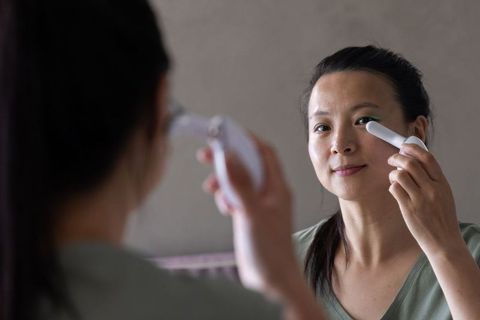 Mature asian woman applying under-eye skincare roller in mirror for gentle anti-aging routine