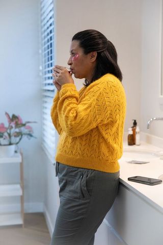 Middle-aged woman sipping from mug at bathroom vanity wearing yellow sweater and under-eye patches