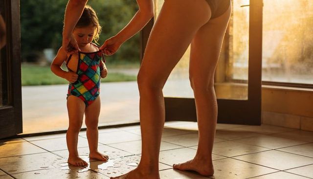 Parent adjusting toddler swimsuit straps on sunlit wet tile by open patio door
