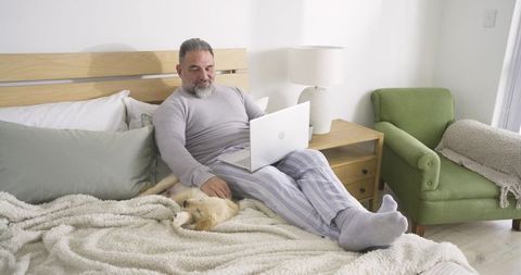 Senior Man Relaxing on Bed with Puppy and Laptop in Cozy Bedroom