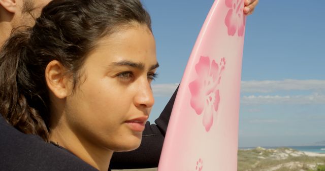 Young biracial woman holds a surfboard at the beach, with copy space. She appears focused and ready for a day of surfing in the sun.