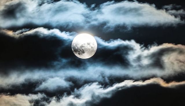 Full moon breaking through silver-blue clouds over dark night sky, dramatic lunar glow