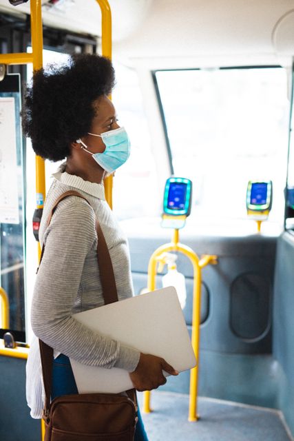 African American businesswoman standing on a bus, wearing a mask and holding a laptop. Ideal for illustrating themes related to commuting, public transportation, business travel, health precautions during the pandemic, and safety measures in public spaces.