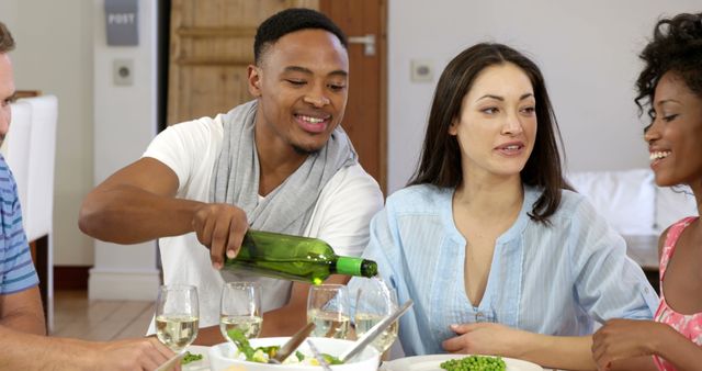 Group of friends gathered at dining table, enjoying food and wine. Young man is pouring wine into a glass while others smile and chat. Perfect for illustrating concepts of friendship, social gatherings, bonding, and home dining experiences.