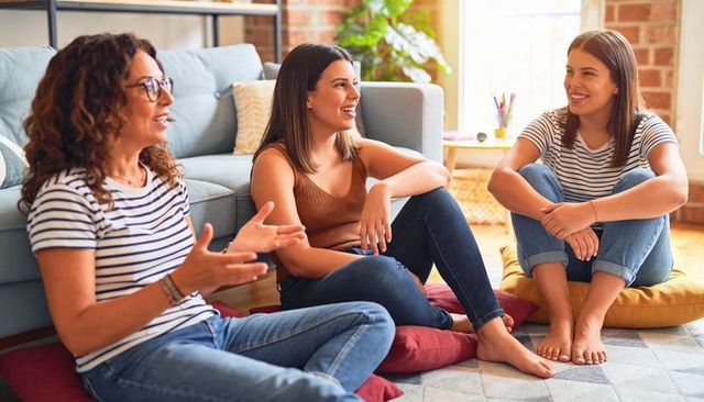 Three women chatting on living room floor cushions, casual cozy friendship conversation