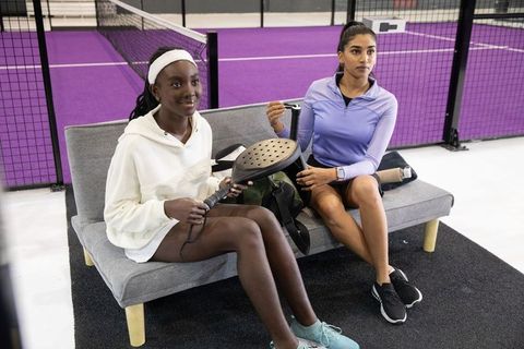 Diverse female teammates sitting at purple padel court holding paddles and smiling