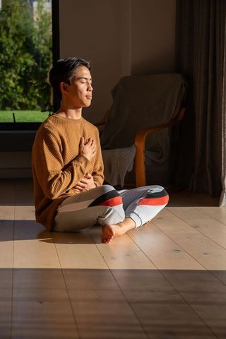 Practicing mindful breathing asian man meditating cross-legged in sunlit cozy living room