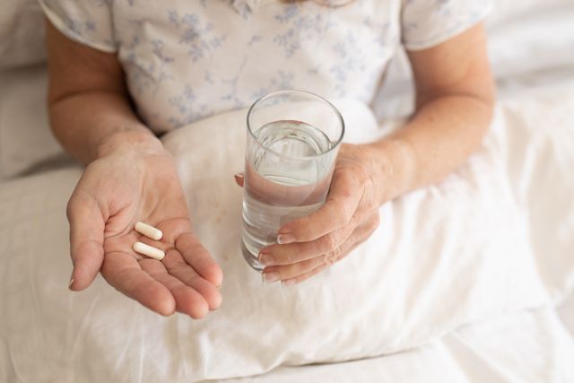 Senior woman holding pills and a glass of water while lying in bed. Ideal for topics related to elderly healthcare, medication, home treatment, and self-care during quarantine. Useful for articles, blogs, and advertisements focusing on health and wellness for seniors, especially during the Covid-19 pandemic.