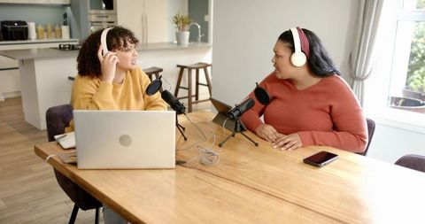 Diverse female cohosts recording home podcast at kitchen table with microphones, laptop