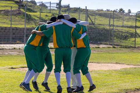 Huddling baseball team linking arms before game on sunny diamond wearing green uniforms