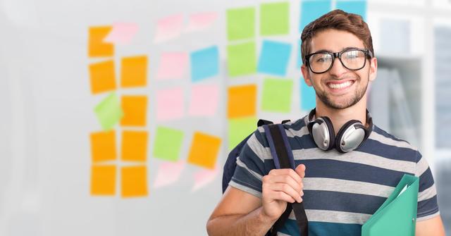 Young professional male with spectacles, holding a backpack and notebooks, smiling confidently in a modern office setting. Multicolored post-it notes on the wall indicate a brainstorming or creative session. Suitable for business, education, or lifestyle content showcasing modern work environments, youth employment, or creative processes.
