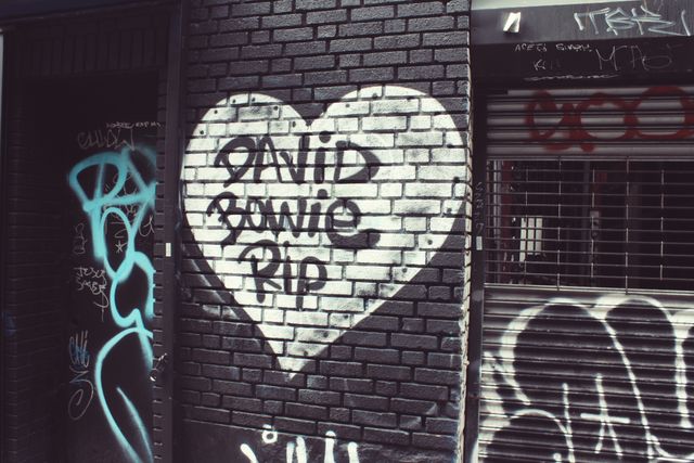 Heart-shaped graffiti with 'David Bowie RIP' on brick wall, symbolizing a tribute or remembrance. Suitable for urban art collections, cultural studies, tributes, or street art enthusiasts. Can be used in articles, blog posts, social media, or designs focusing on urban art, tributes to artists, and expressions of public mourning.