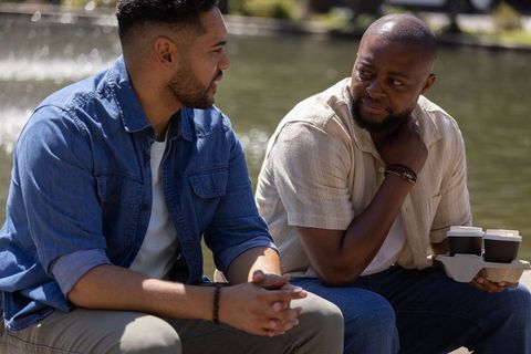 Two african american men talking on bench by fountain sharing coffee and conversation