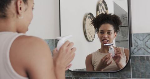 African american woman holding facial grooming device looking in mirror for skincare