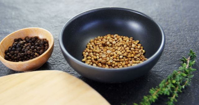 A black bowl filled with coriander seeds is placed on a dark surface, with copy space. Nearby, a wooden bowl contains black peppercorns and a sprig of thyme adds a touch of greenery, suggesting a setting for culinary preparation or recipe ingredients.
