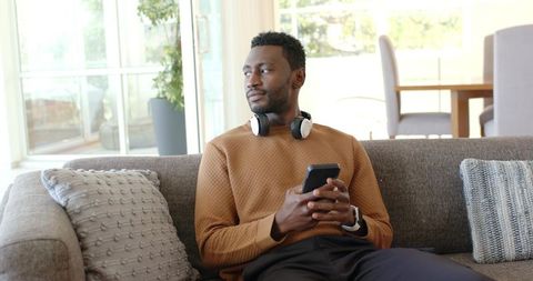 Man sitting on gray sofa wearing camel sweater and headphones holding smartphone