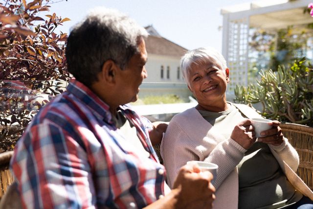 Senior African American couple sitting on a terrace, enjoying coffee together. They are smiling and appear relaxed, suggesting a peaceful retirement lifestyle. This image can be used for topics related to retirement, elderly care, self-isolation during the COVID-19 pandemic, and promoting a healthy, relaxed lifestyle for seniors.