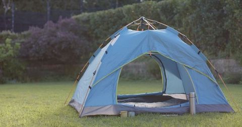 The camping tent sits in a lush green backyard, offering an inviting retreat in nature. Sunlight illuminates the scene, emphasizing relaxation potential. Accompanied by a thermos and cups, the setup is perfect for planning staycations or showcasing leisurely outdoor lifestyles. Suitable for advertisements or articles about camping gear, backyard activities, or embracing nature at home.