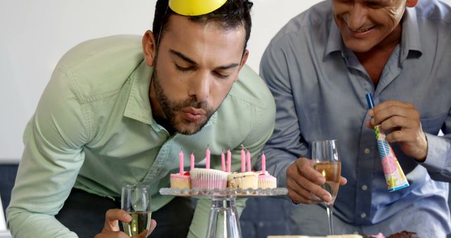 A young Caucasian man blows out candles on a birthday cake, with copy space. An older man, his colleague or friend, watches with a party blower, sharing in the celebration.