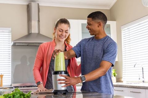 Preparing green smoothie together in athleticwear at modern kitchen island wearing smartwatches