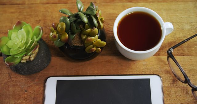 A cup of tea, a tablet, eyeglasses, and a small potted plant are neatly arranged on a wooden surface, with copy space. This setup suggests a relaxed work or reading environment, indicating a break or a moment of leisure.