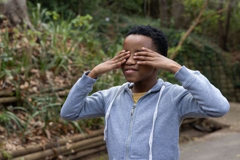 African american teen covering eyes on forest trail wearing gray hoodie and yellow tee