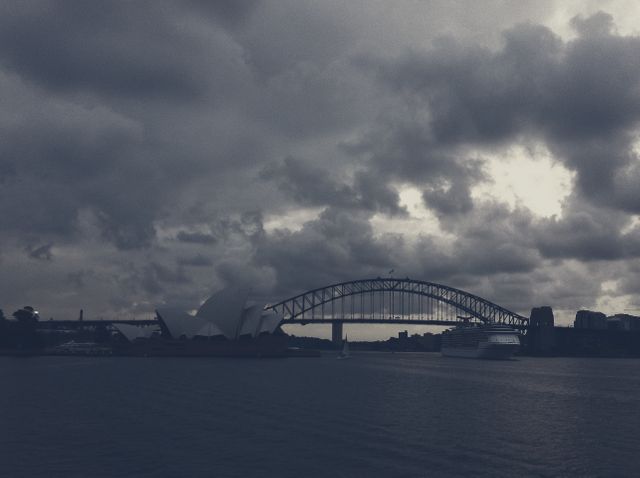Image showcasing the dramatic view of Sydney's Harbour Bridge and Opera House with dark clouds at dusk. Can be used in travel brochures, articles about Sydney or Australia, weather blogs, or urban architecture studies. Highlights the iconic landmarks under a stormy sky, emphasizing the moody atmosphere.