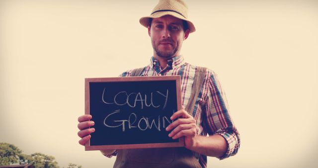 Farmer holding chalkboard sign advertising locally grown produce, wearing hat and plaid shirt in rural setting. Perfect for promoting farmers' markets, sustainable agriculture, organic food initiatives, and community-supported agriculture programs.