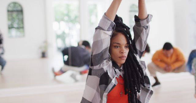 Young woman with dreadlocks in plaid shirt stretching with focus in dance studio, others in background. Suitable for themes related to fitness, mindfulness, contemporary dance, empowerment, diversity, and urban fashion.