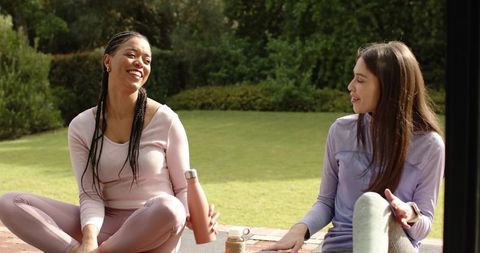 Laughing friends relaxing after outdoor workout on patio, chatting in pastel activewear