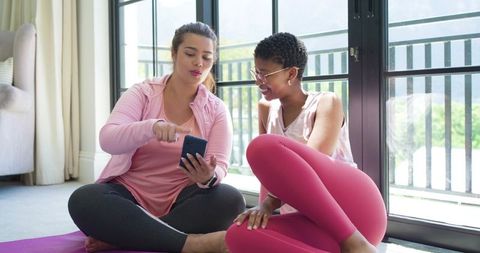 African american woman and friend sharing smartphone on yoga mat by sunlit balcony