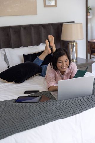 Asian woman relaxing on bed using laptop and notepad for remote work and study